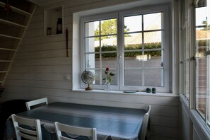 A dining area with a table and chairs, a window with a view of the outside, and a shelf with items. - Charmante Maison Parfaitement Équipée Pour 4 Pers. Emplacement Ideal (Barneville Carteret)