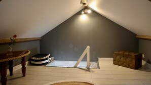 Une pièce avec un plafond en pente, une table ronde surmontée d’un vase, un panier pour animal de compagnie, une chaise blanche et une boîte de rangement.