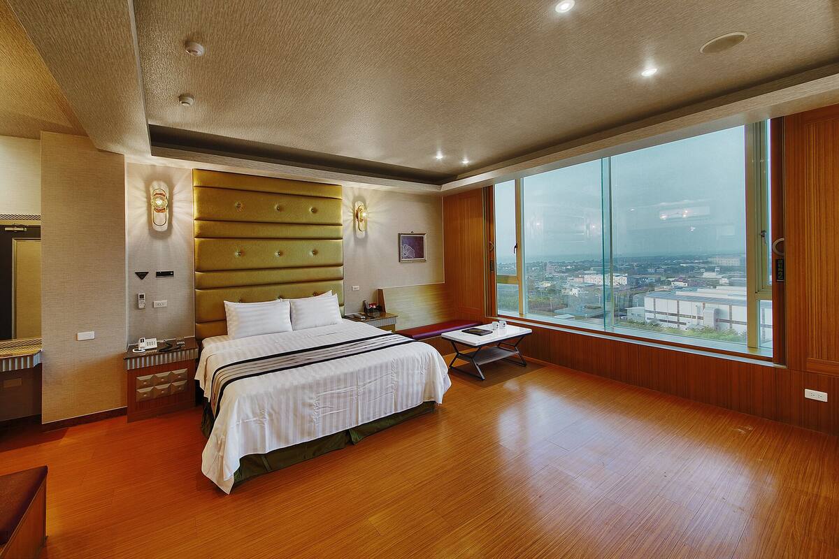 Una habitación de hotel amplia con una cama grande, suelo de madera, vistas de la ciudad a través de amplias ventanas y un diseño de techo moderno.