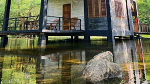 Una casa de madera con balcón, ubicada sobre una masa de agua, rodeada de árboles.