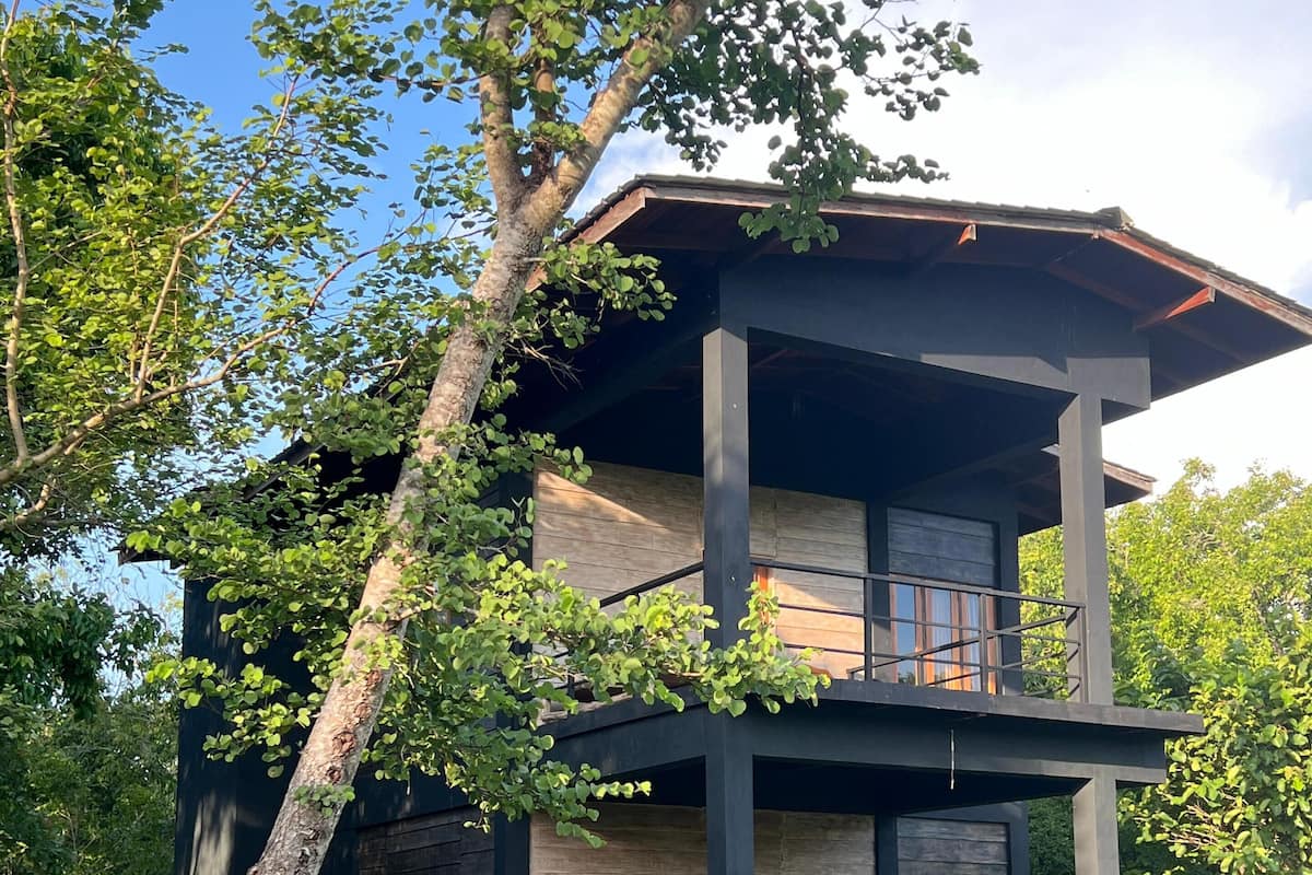 A two-story cabin with a balcony, surrounded by trees and a small body of water.