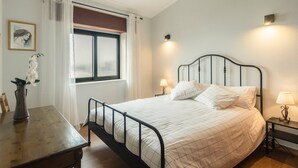 A bedroom with a bed, bedside lamps, a window with curtains, a desk with a vase of flowers, and a framed picture on the wall. - Casa Curva do Rio (Melres)