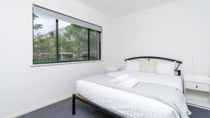 Quarto de hotel com uma janela grande, cama com roupa branca, mesa de cabeceira e criado-mudo.