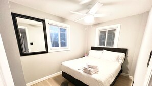 A bedroom with a bed, two windows with blinds, a ceiling fan, and a large mirror. - SeaSide Serenity - Cozy Retreat on Bay Drive (Kill Devil Hills)