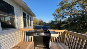 A barbecue grill on a wooden deck with a house in the background. - SeaSide Serenity - Cozy Retreat on Bay Drive (Kill Devil Hills)