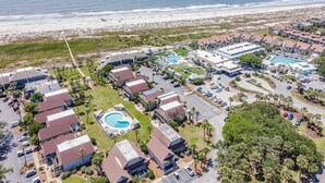Eine Luftaufnahme eines Küstenresorts mit mehreren bungalowsähnlichen Gebäuden, einem zentral gelegenen Schwimmbad und einem Strand im Hintergrund.