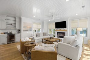 A spacious living room with a fireplace, a television mounted on the wall, and a kitchen area with white cabinetry.