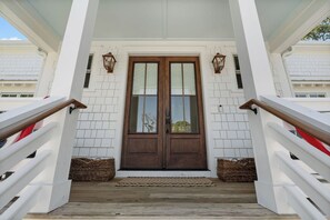 A wooden front door with glass panels, white brick walls, and a wooden porch with white railings.