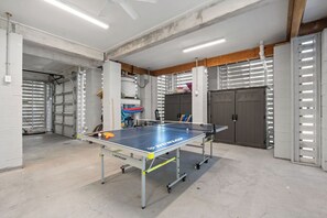 Une table de ping-pong dans une pièce dont le sol est en béton, qui est équipée d’un ventilateur de plafond et dont la porte est munie d’un volet métallique.