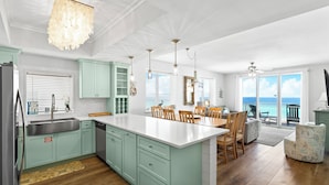 A spacious kitchen with light green cabinets, a white island, and a dining area with wooden chairs. - Majestic Sun 501B - Updated Corner-Unit, Direct Gulf Views, Unmatched Amenities! (Miramar Beach)