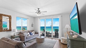 A spacious living room with a large TV, a ceiling fan, and a view of the ocean through the sliding glass doors. - Majestic Sun 501B - Updated Corner-Unit, Direct Gulf Views, Unmatched Amenities! (Miramar Beach)