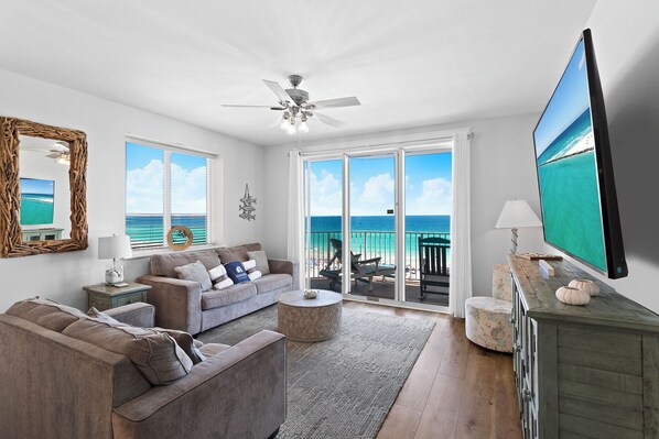 A spacious living room with a large TV, a ceiling fan, and a view of the ocean through the sliding glass doors.
