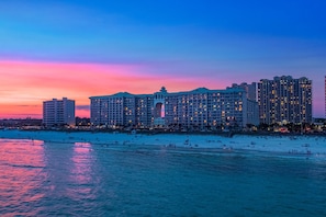 A large hotel with multiple floors and a central courtyard, located near a beach with people on the sand and water.