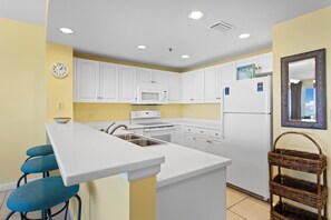 A compact kitchen with white cabinets, a microwave, a stove, and a refrigerator. There is a clock on the wall and a wicker basket on the right.