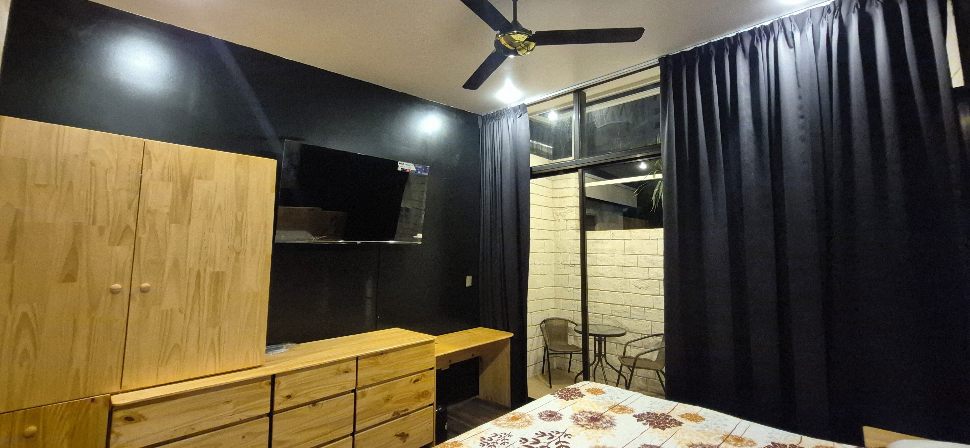 A bedroom with a bed, a wooden dresser, a TV mounted on the wall, a ceiling fan, and a window with curtains.