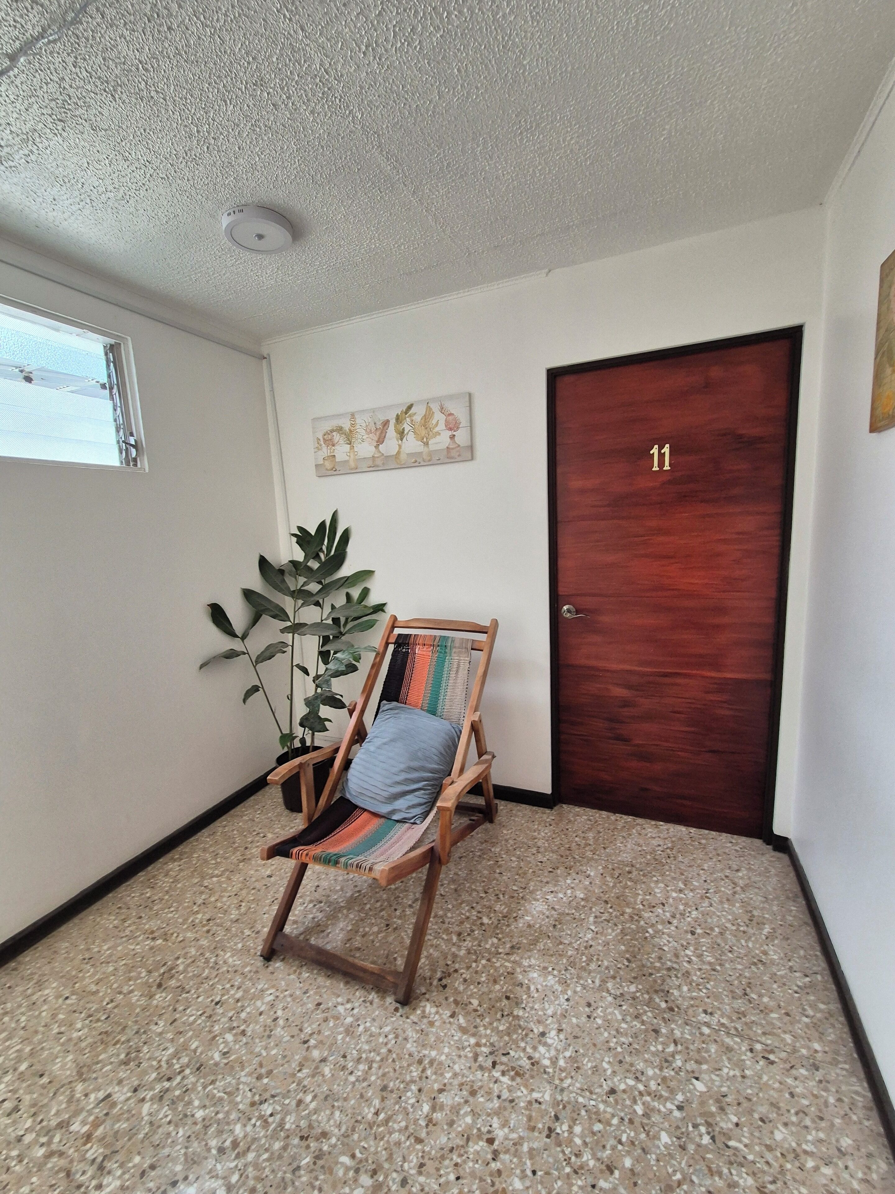 A room with a wooden door marked with the number 11, a potted plant, a wooden chair with a cushion, and a ceiling with a smoke detector.