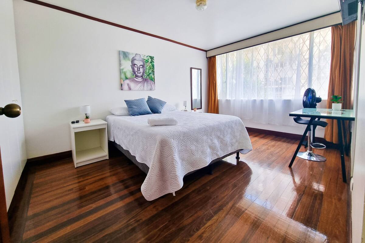 A hotel room with a bed, bedside table, lamp, and a desk with a fan.