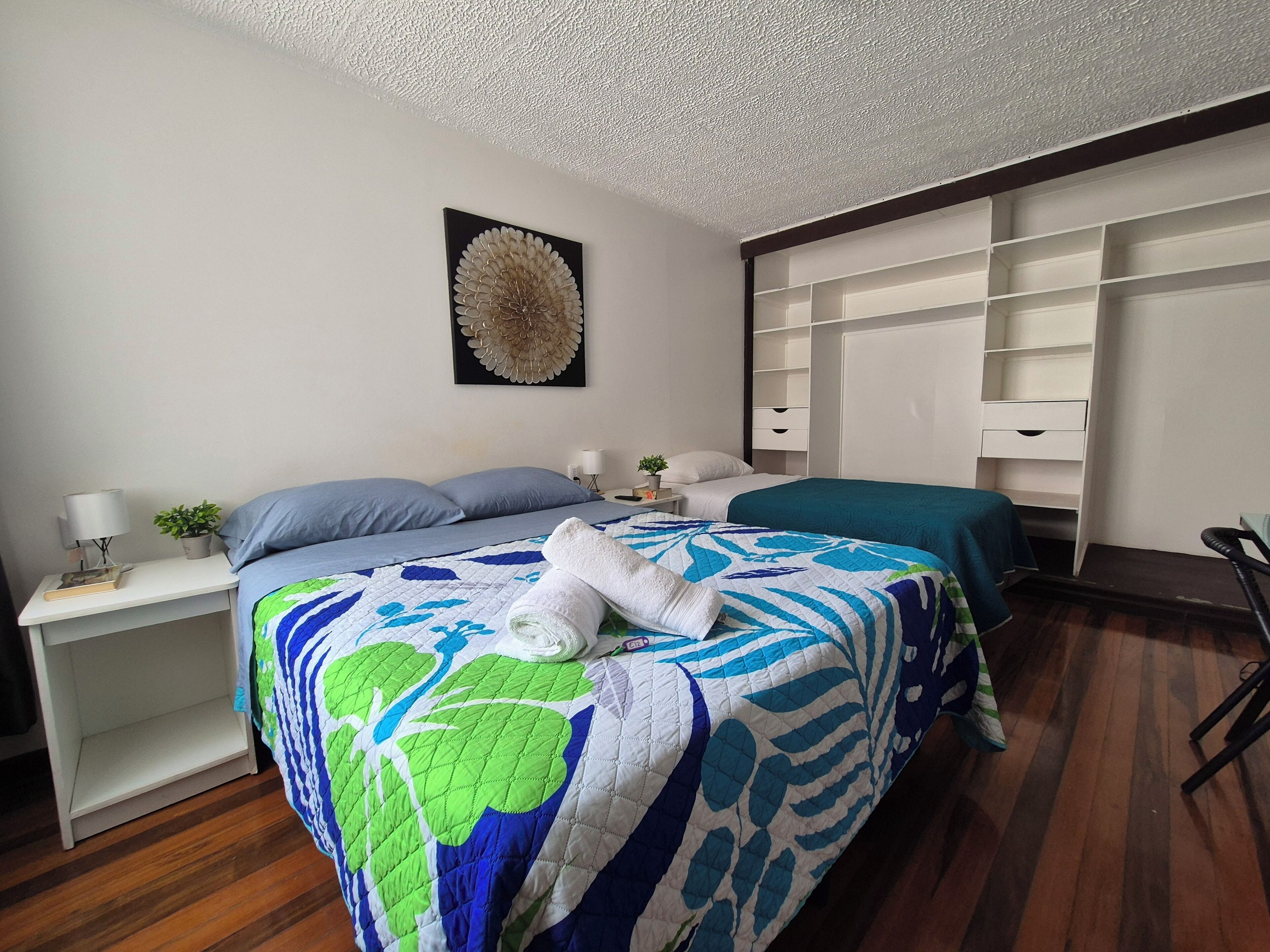 Quarto bem arrumado com uma cama grande, uma mesa de cabeceira com abajur, uma pequena planta e um objeto decorativo na parede.