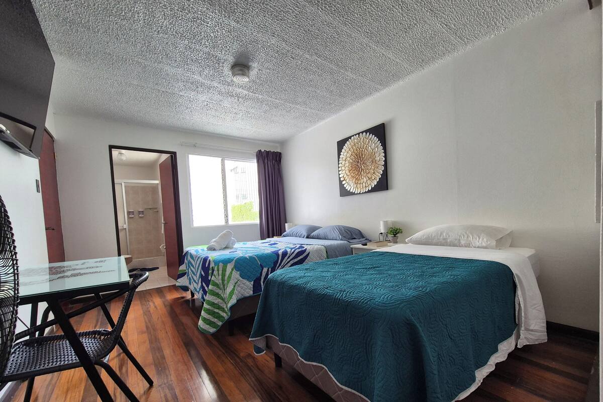 A hotel room with two beds, a wooden floor, a ceiling fan, a window with curtains, and a wall-mounted artwork.