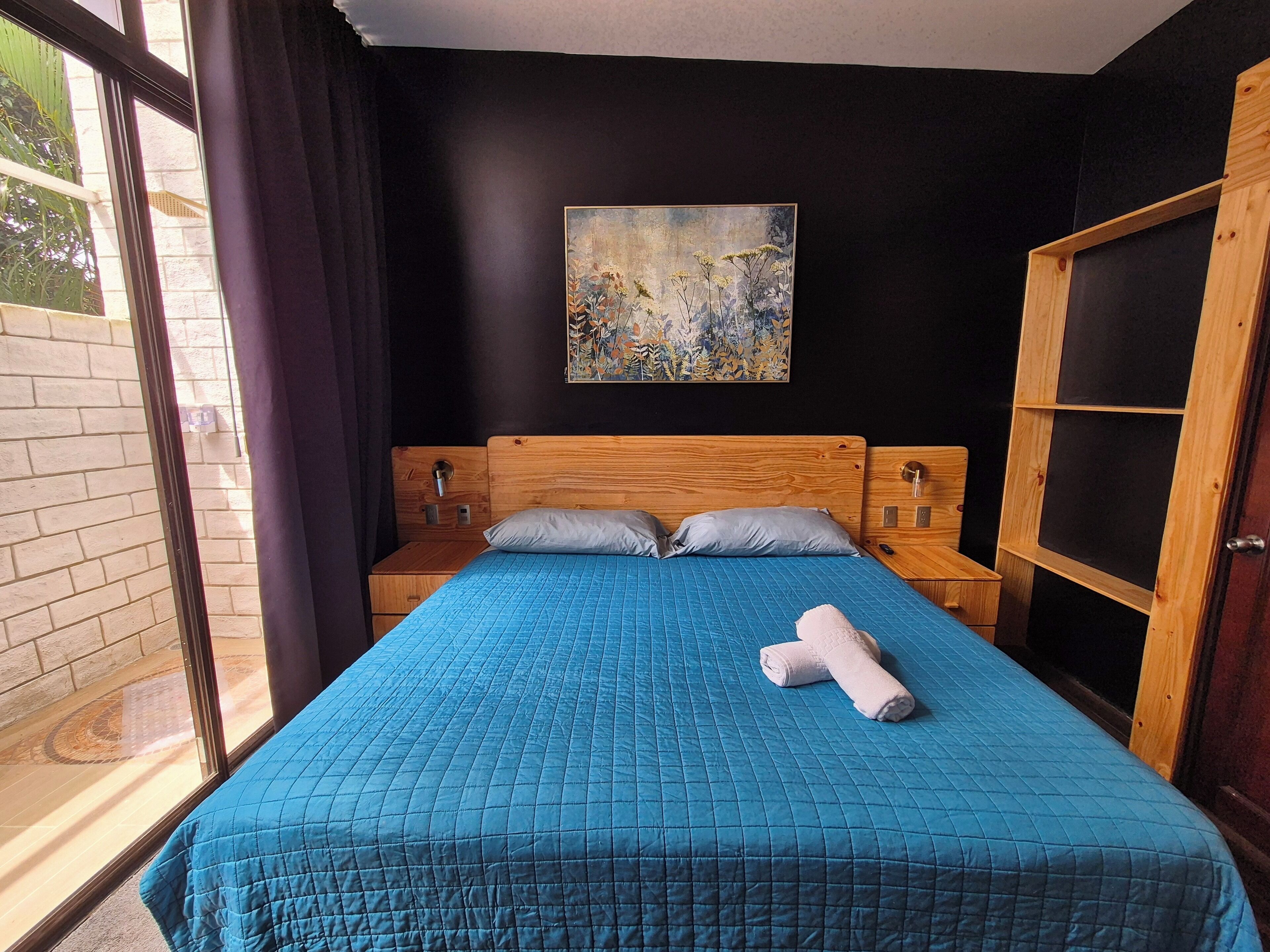 A bedroom with a large bed, two bedside tables, a painting on the wall, and a view through a window.