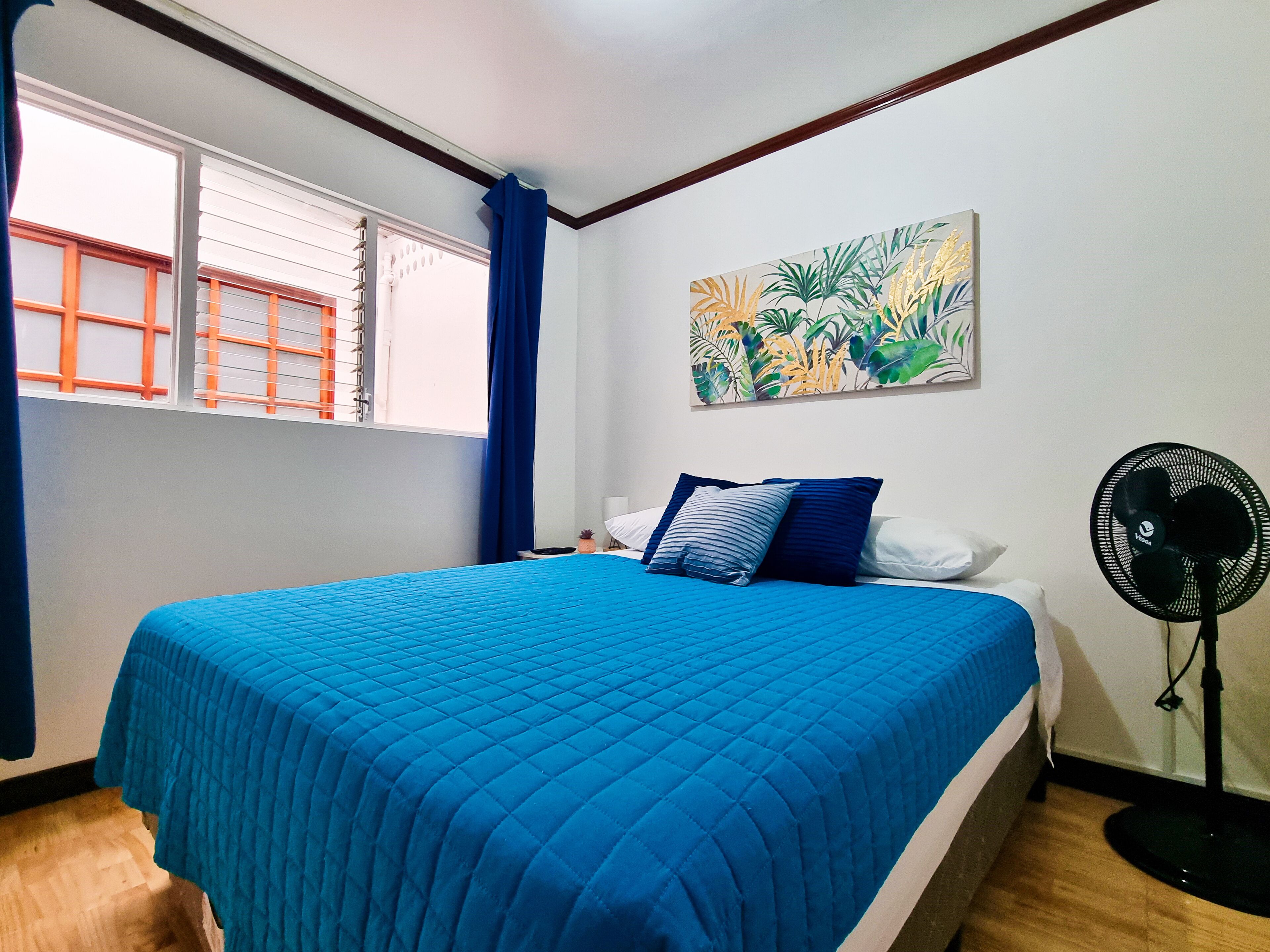 Un dormitorio con cama, ventana con persianas, un cuadro y un ventilador de pie.