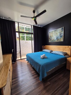 A bedroom with a bed, a ceiling fan, a wooden nightstand, a wooden bench, and a view of greenery outside. - Sabana Studios (San José)