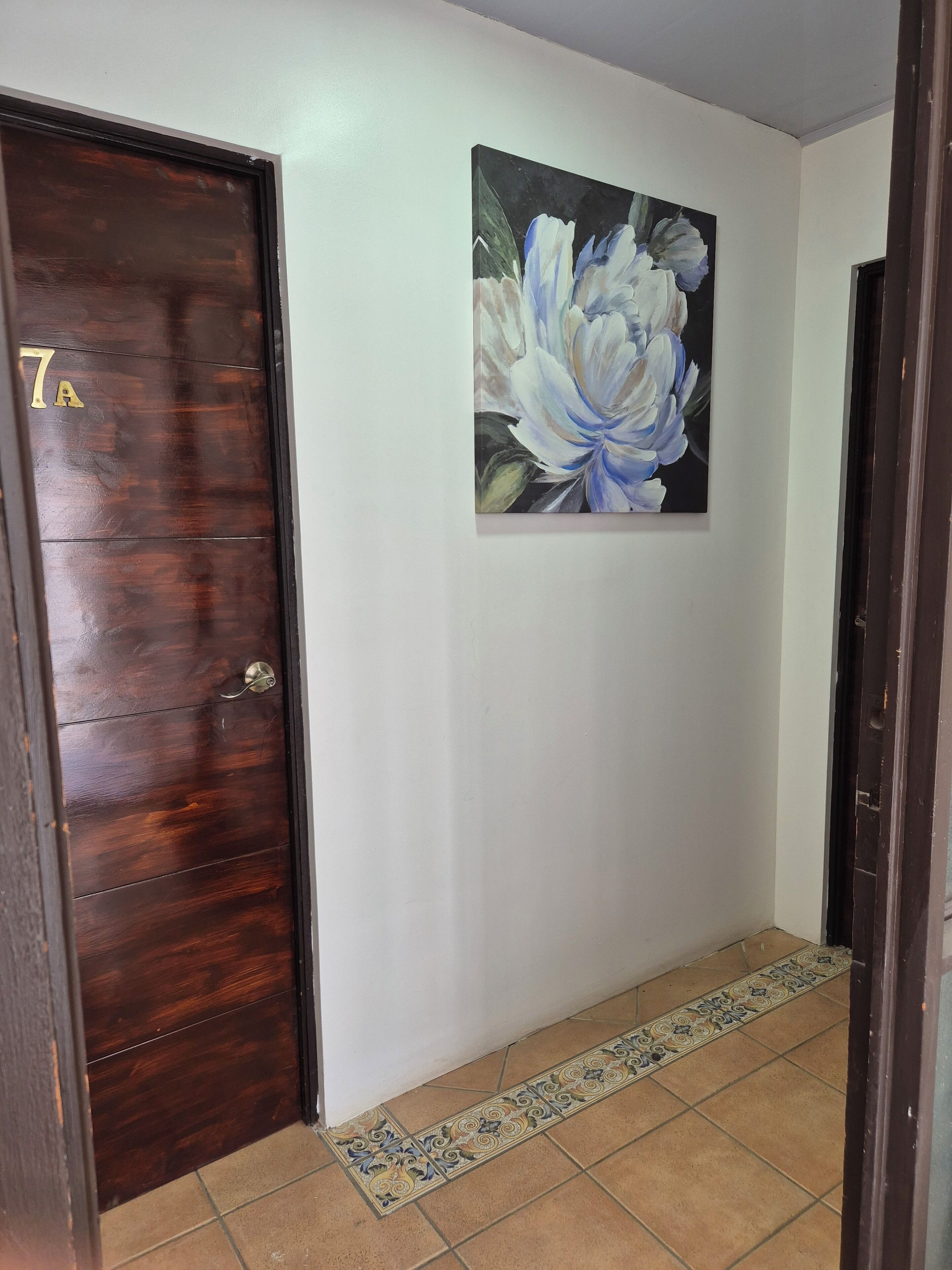 A hallway with a wooden door marked "7A", a tiled floor, and a floral painting on the wall.