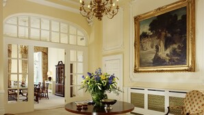 A grand room with a round table, a chandelier, a large painting, and a view of a dining area. - Cheval Hyde Park Gate (London)