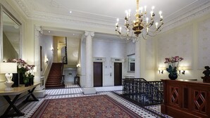 A grand lobby with a large ornate chandelier, a patterned carpet, and a staircase leading to an upper floor. - Cheval Hyde Park Gate (London)