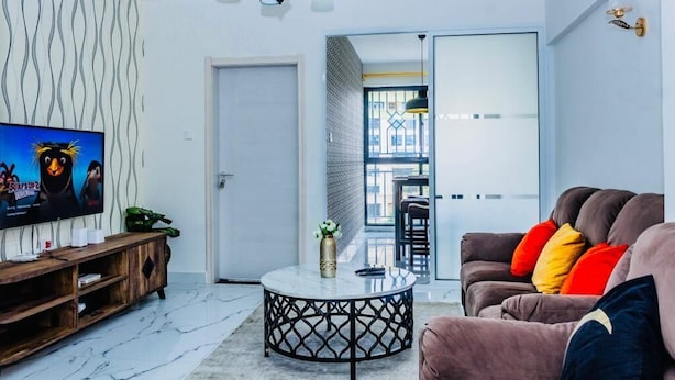 A modern living room with a TV, a round coffee table, a sofa with colorful cushions, a wooden entertainment unit, and a patterned wall. - Lux Suites Micasa Sandalwood Apartments (Nairobi)