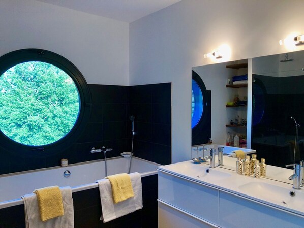 A modern bathroom with two sinks, a large mirror, and a circular window with a view of greenery.