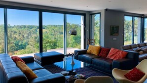 Ein modernes Wohnzimmer mit einem großen Glasfenster, Ledersofas, einem Glastisch und Blick auf eine bewaldete Landschaft.