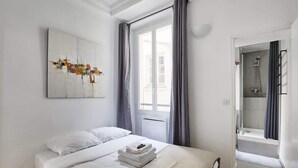 A hotel room with a bed, a window with curtains, a shower area, and a piece of abstract art on the wall. - Apartment  Center of Paris (Paris)