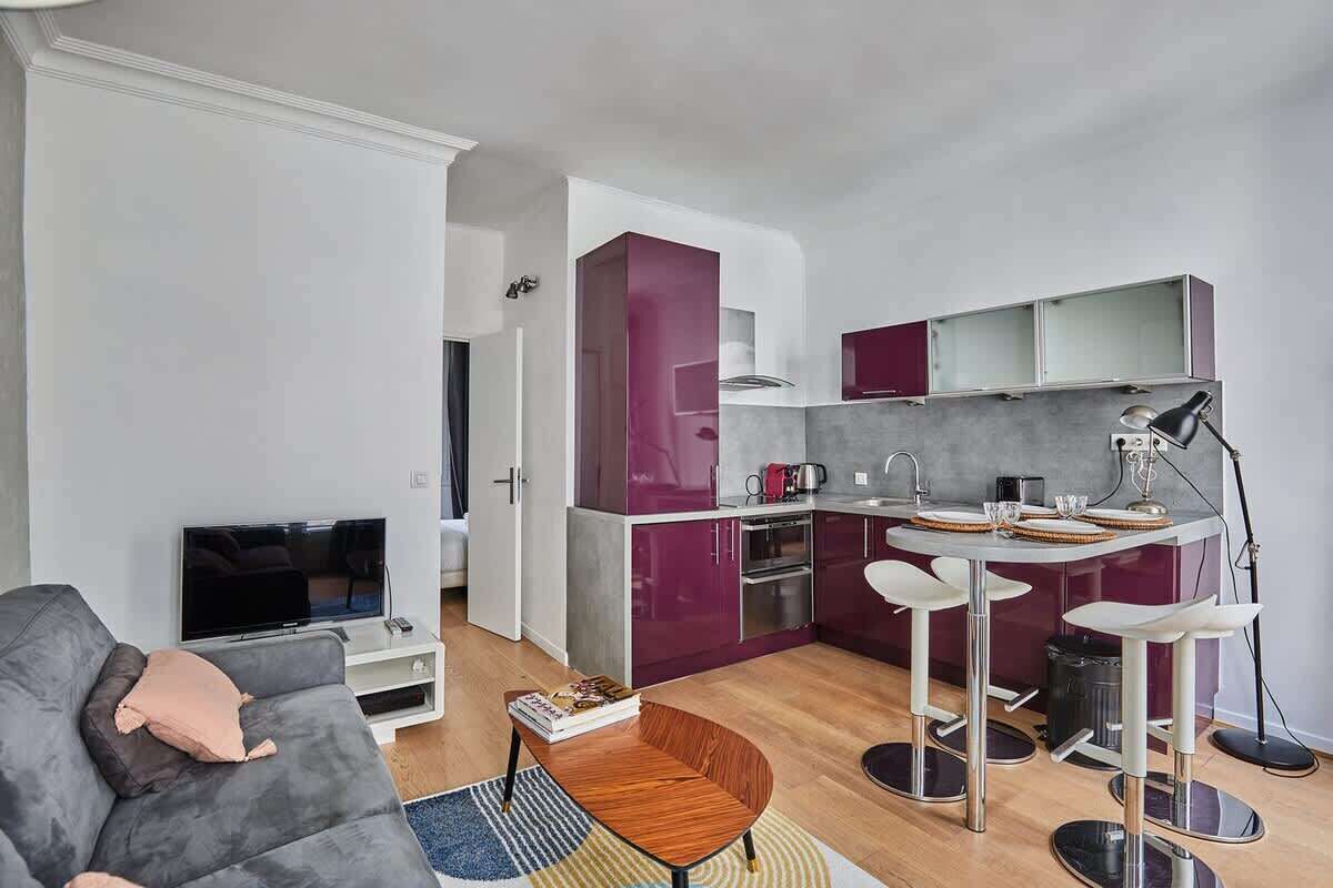 Een compacte leefruimte met een grijze bank, een klein houten bijzettafeltje, een televisie, een keukenhoek met een eettafel en barstoelen, en een kitchenette met paarse kastjes.