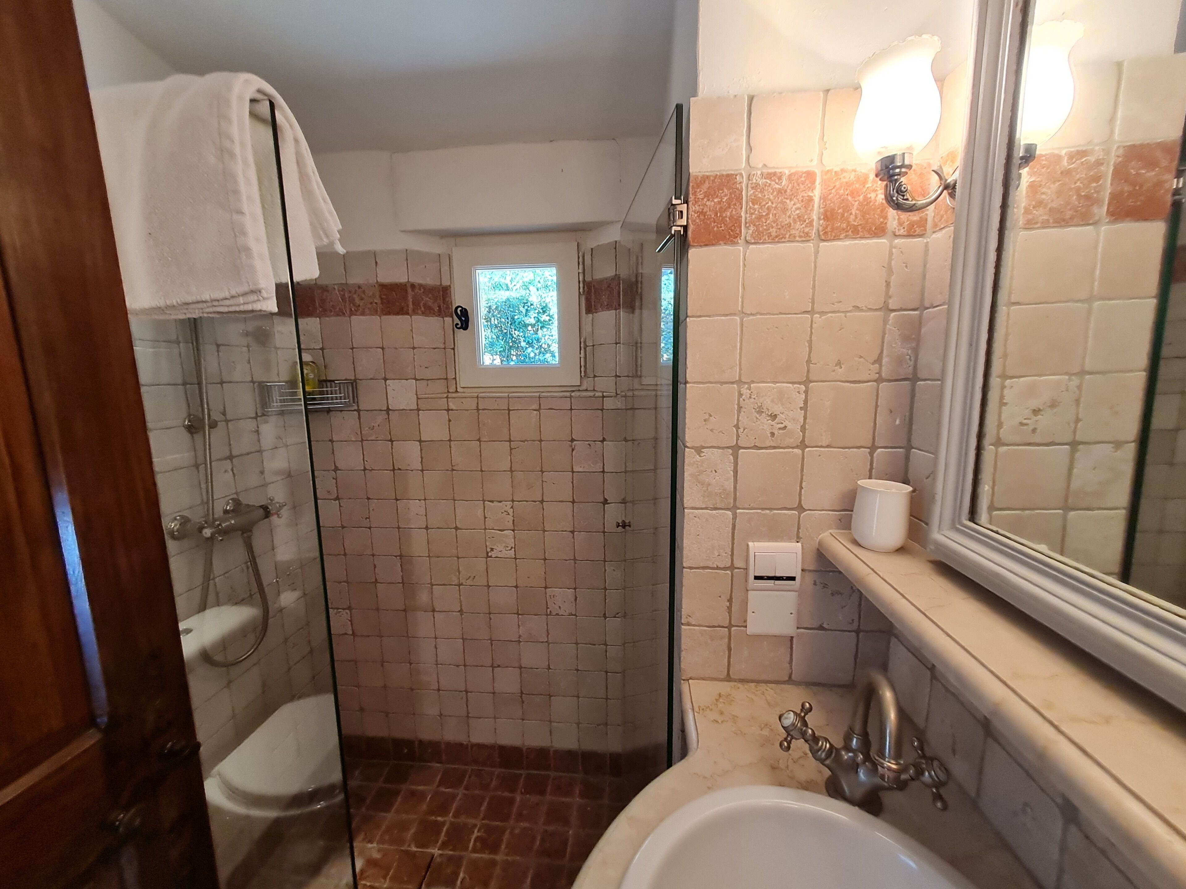 A bathroom with a glass shower enclosure, a toilet, a sink with a mirror, and a small window.