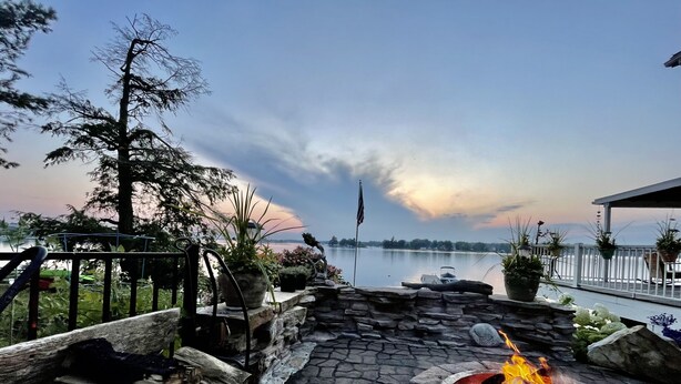 Une terrasse en pierre avec un foyer, des plantes en pot et une vue sur un lac, avec un bateau et des montagnes à l’horizon.