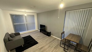 A modern living room with a grey sofa, a black rug, a wooden dining table with chairs, a TV on a stand, and large windows with blinds.