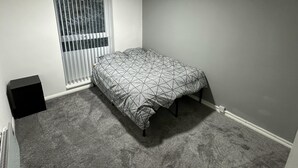 A bedroom with a single bed, a window with blinds, a carpeted floor, and a small black speaker.