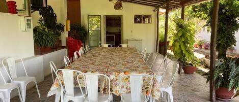 A covered dining area with a long table and white chairs, surrounded by potted plants and a stone-tiled floor.