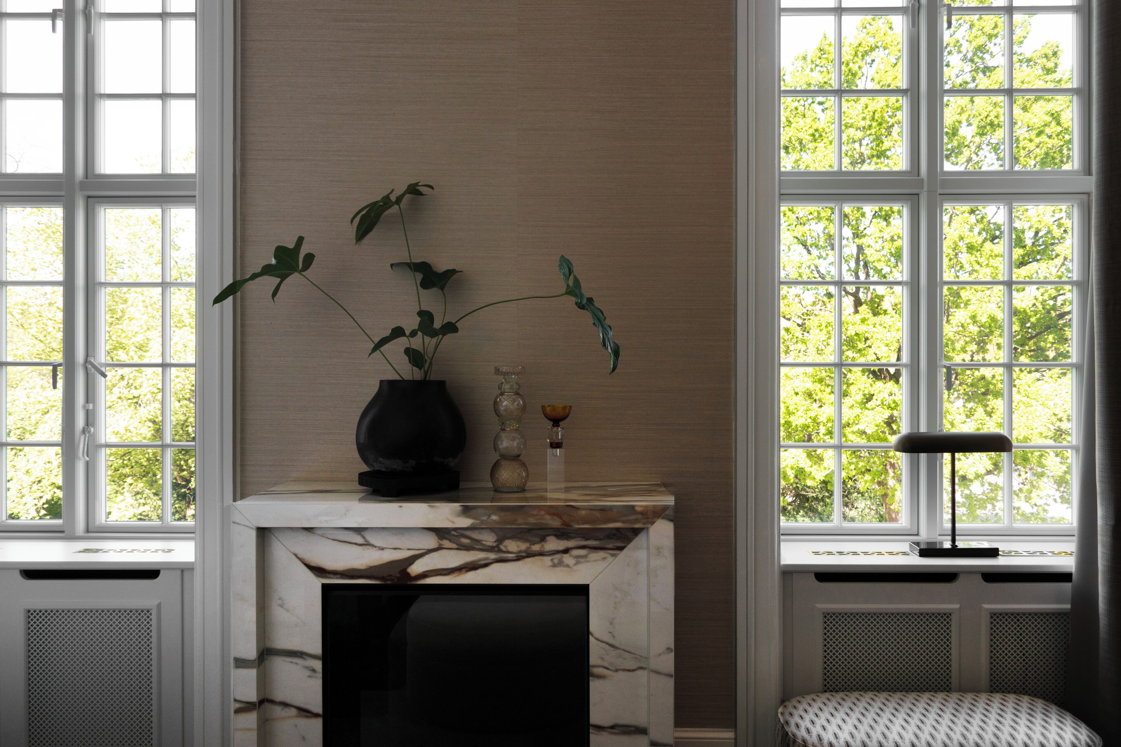 A room with a marble fireplace, a vase with a plant, and a window with a view of trees.