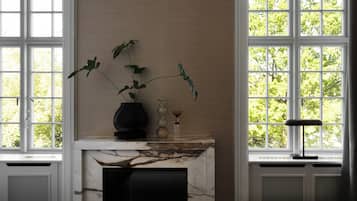 A room with a marble fireplace, a vase with a plant, and a window with a view of trees.