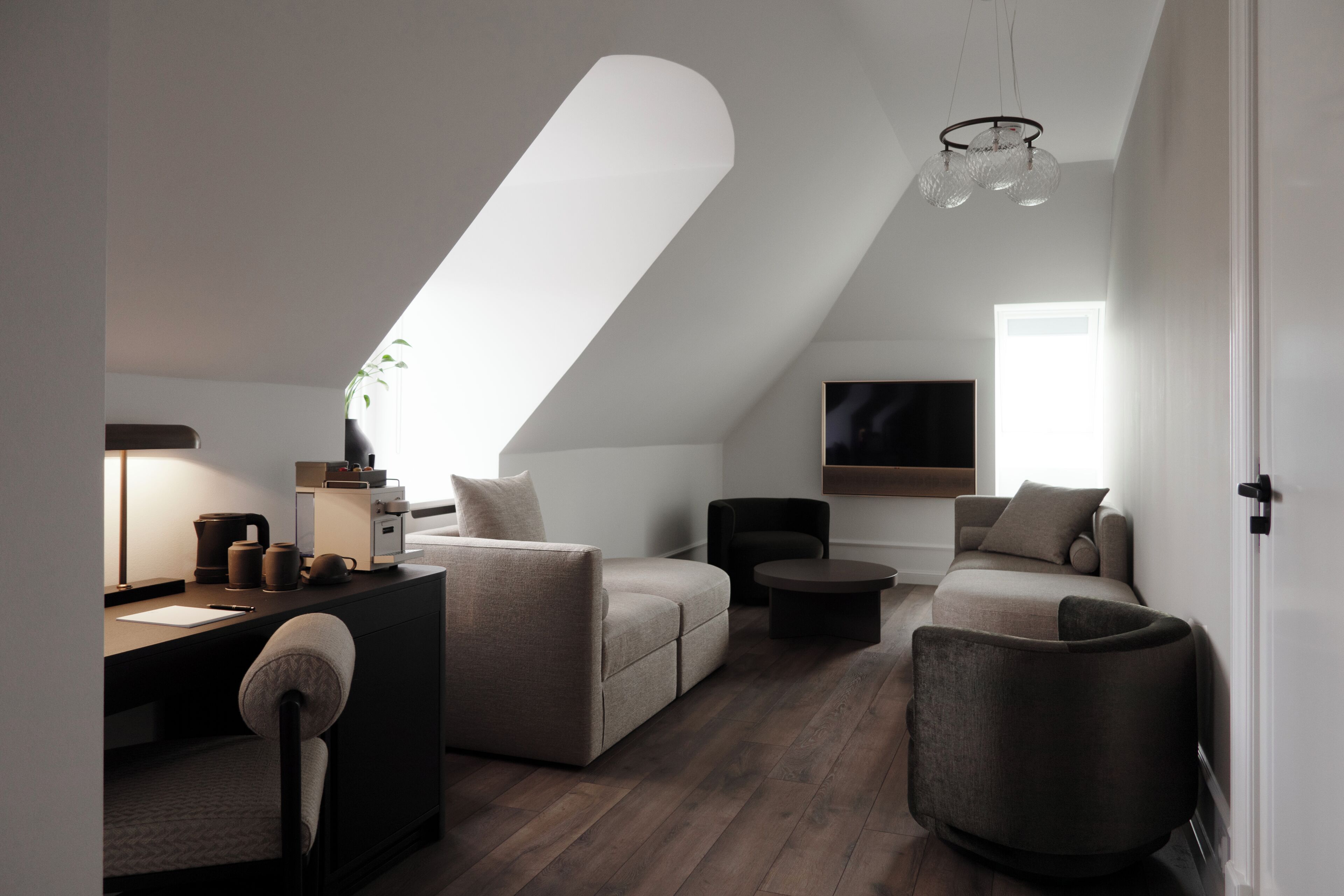 A modern loft bedroom with a sloped ceiling, a large window, a flat-screen TV, and a cozy seating area with a desk and chair.