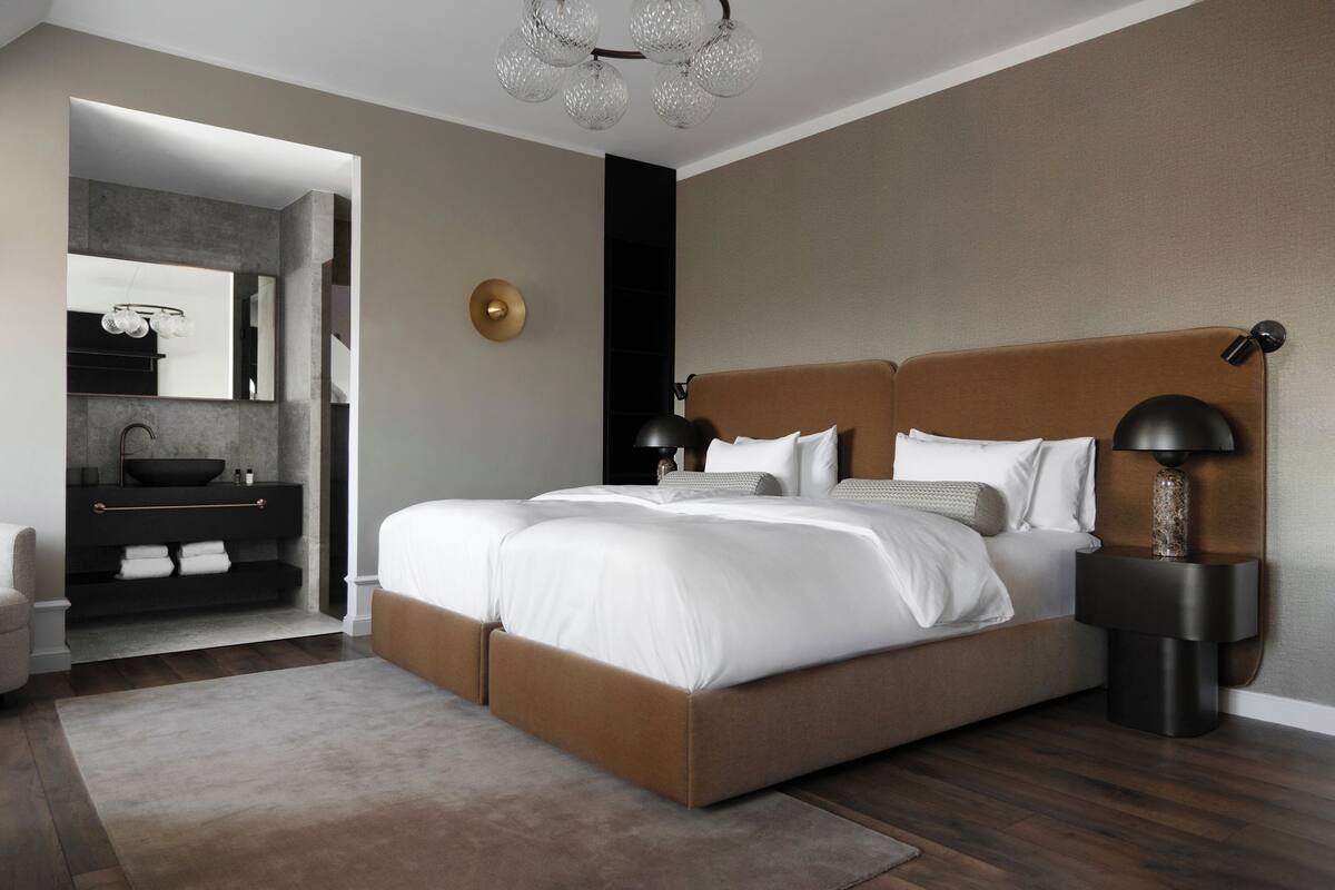 Un dormitorio moderno con una cama grande, lámparas de noche, una mesita y un baño visible a través de un umbral abierto.