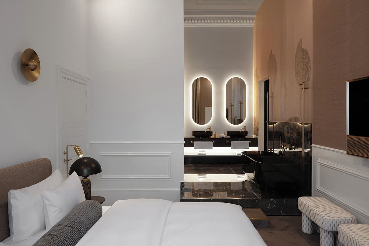 Una habitación de hotel moderna con una cama grande, una lámpara de noche, una mesita de tocador con espejo y una bañera.