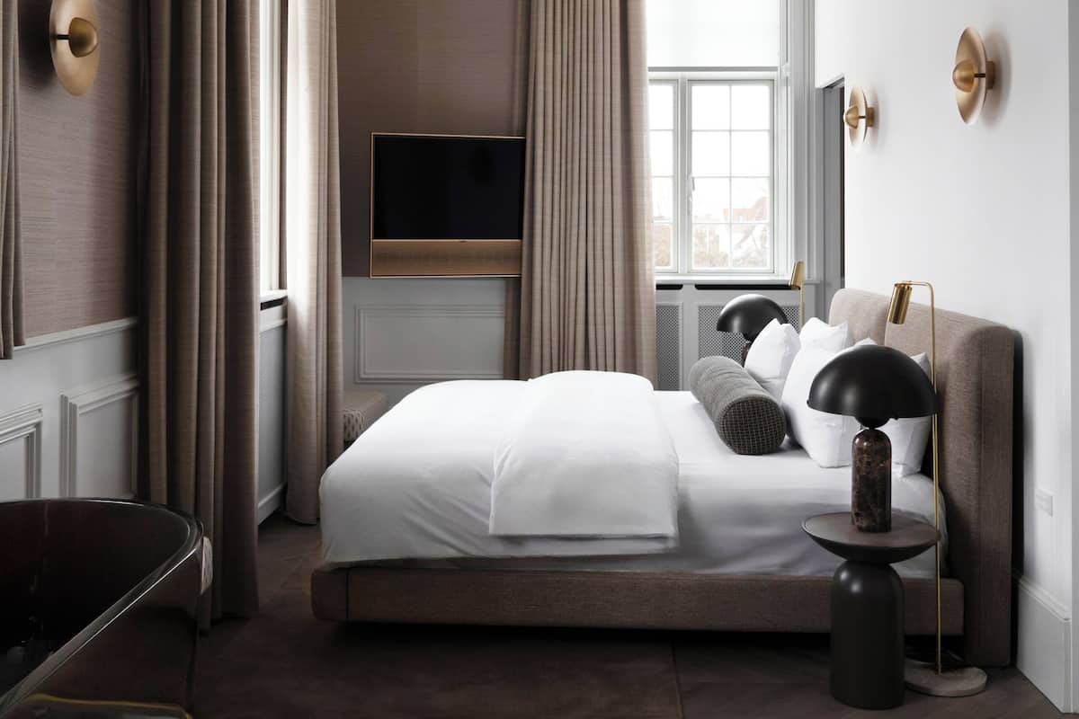 Una habitación de hotel moderna con una cama grande, un televisor de pantalla plana, una mesita de noche con lámpara y vistas a la ciudad desde la ventana.