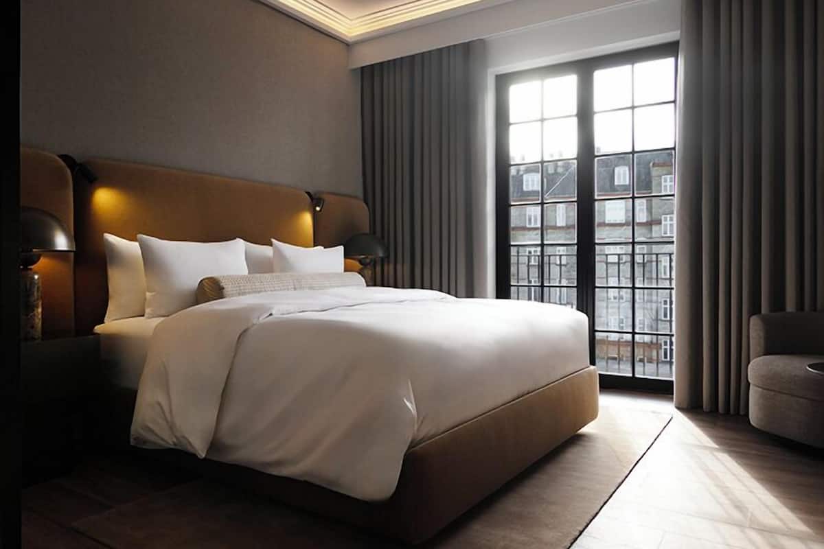 Habitación de hotel moderna con una cama grande, una mesita de noche, una lámpara y vistas a la ciudad a través de amplias ventanas.