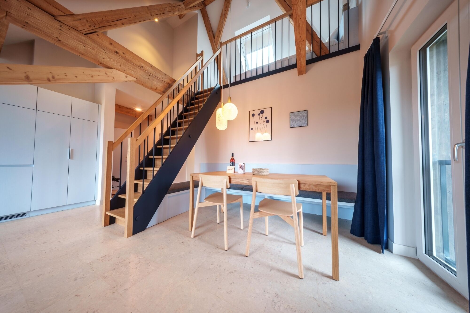 A spacious room with a wooden staircase, a dining area with a table and chairs, and a large window with curtains.
