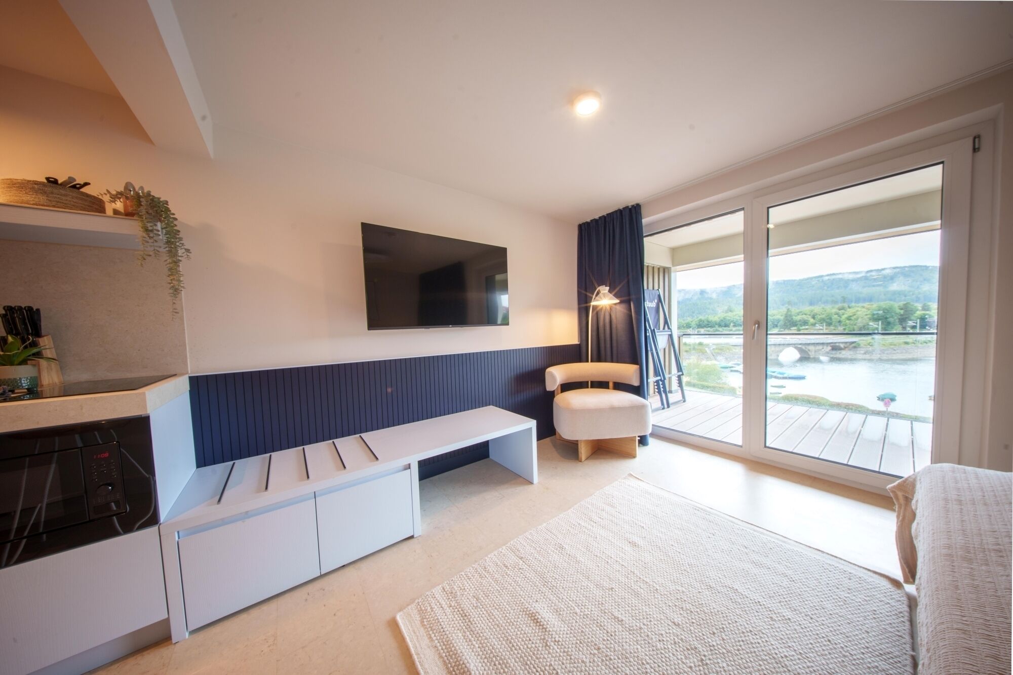 A modern living room with a flat-screen TV, a bench, and a large window offering a view of a river and hills.