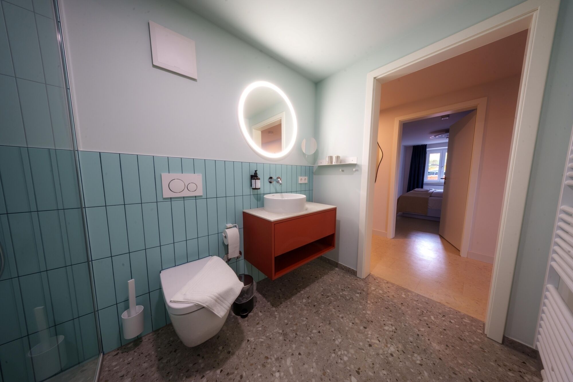 Une salle de bain avec des toilettes blanches, un lavabo et un miroir surmonté d’une lampe circulaire. Une serviette est posée sur les toilettes, et les murs sont recouverts de carreaux de couleur bleu clair.