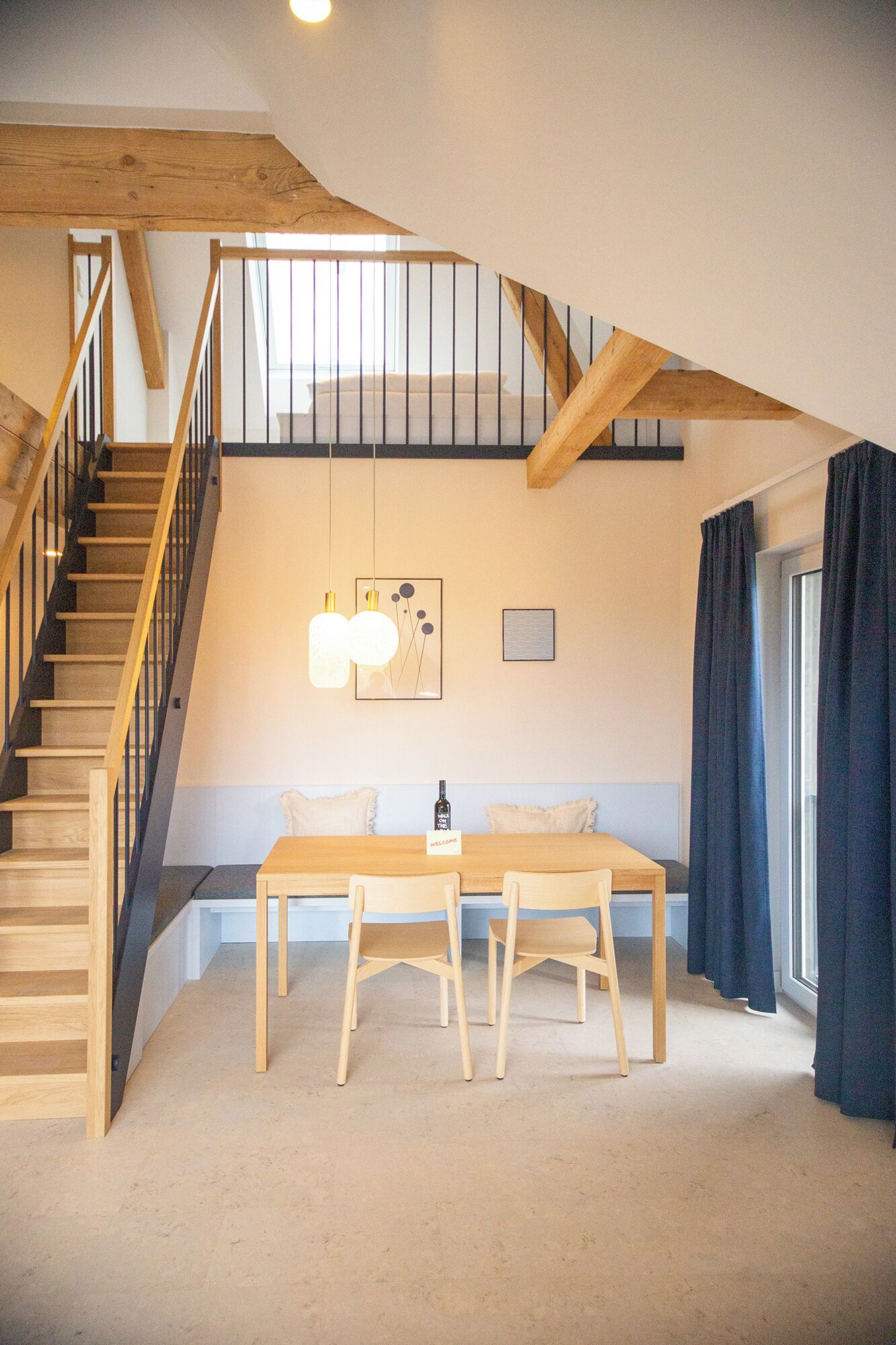 A cozy room with a wooden staircase, a dining table with chairs, a sofa, and a large window with curtains.